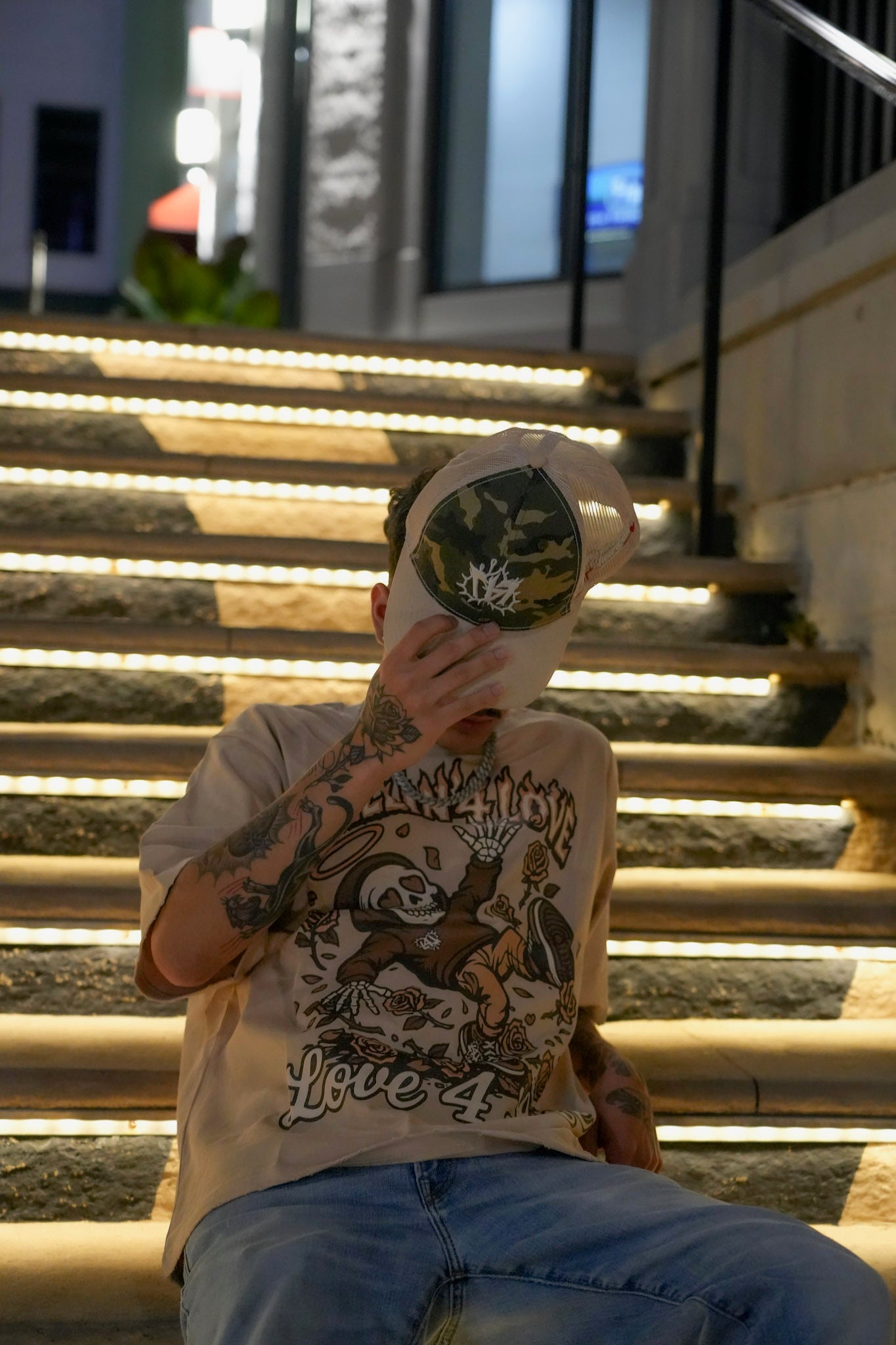 A person sitting on illuminated outdoor stairs at night, wearing a beige graphic t-shirt, blue jeans, and a camouflage Love 4 Life cap held up to block their face.