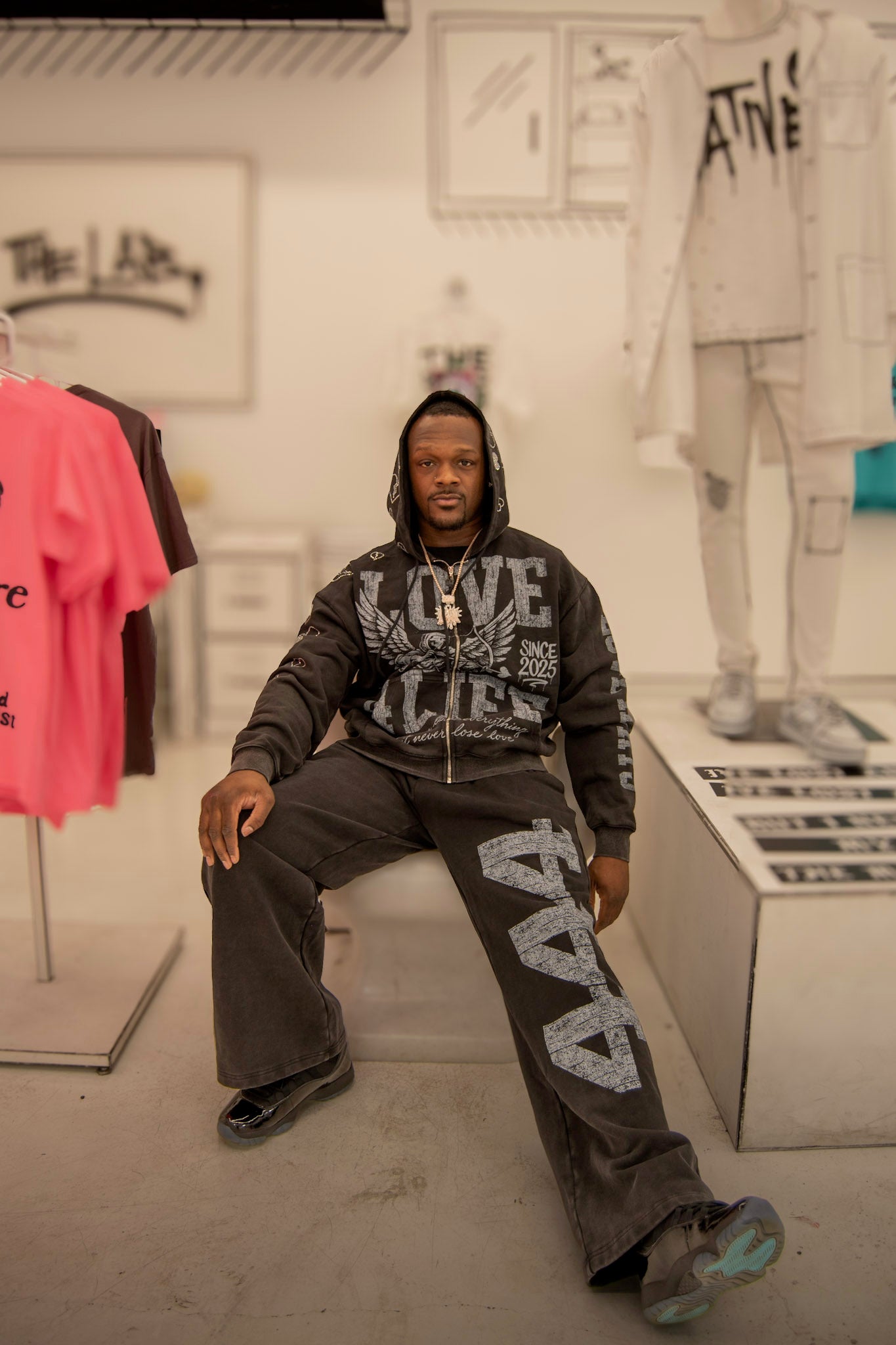 Man sitting in a store wearing a hoodie with text, surrounded by clothing and displays. Love 4 Life Clothing Brand